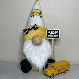 Może przedstawiać: Ż&oacute;łty krasnal w białym kapeluszu z wzorem szkolnego autobusu i czarno-białą kokardką. Krasnal trzyma znak z napisem "School Bus Drivers Gnome Best!" i przed nim stoi ż&oacute;łty, zabawkowy autobus szkolny.