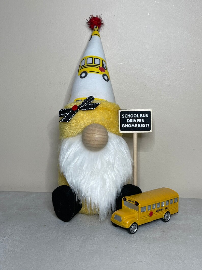 Może przedstawiać: Ż&oacute;łty krasnal w białym kapeluszu z wzorem szkolnego autobusu i czarno-białą kokardką. Krasnal trzyma znak z napisem "School Bus Drivers Gnome Best!" i przed nim stoi ż&oacute;łty, zabawkowy autobus szkolny.