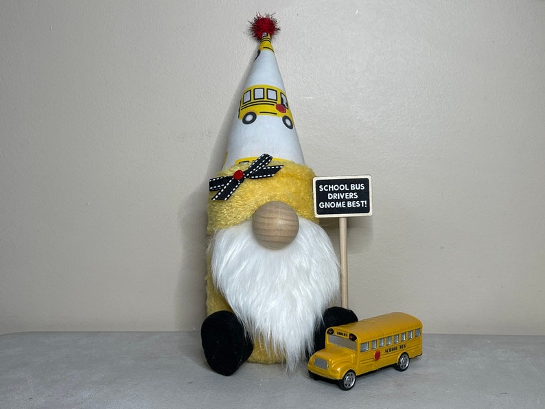 Może przedstawiać: Ż&oacute;łty krasnal z białą brodą i czerwono-białym kapeluszem z wzorem szkolnego autobusu. Krasnal trzyma znak z napisem "School Bus Drivers Gnome Best!" i przed nim stoi ż&oacute;łty, zabawkowy autobus szkolny.