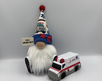 May include: A whimsical gnome with a white beard, blue hat, and red bow tie, holding a sign that reads "E.M.S. Gnomes Best!" Next to it is a toy ambulance with red and white details.