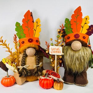 Puede incluir: Dos gnomos de Acción de Gracias con sombreros de fieltro marrón con plumas naranja, amarillo y verde. Los gnomos sostienen un cartel que dice "Give Thanks".