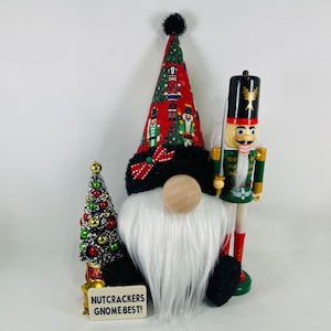 May include: A Christmas gnome with a red and green hat featuring a nutcracker pattern. The gnome has a white beard and a wooden nose. A small Christmas tree and a nutcracker stand beside the gnome. The sign reads "Nutcrackers Gnome Best!"