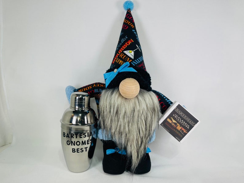 Pu&ograve; includere: Un gnomo con un cappello nero e blu con una stampa di cocktail e un fiocco blu e bianco. Il gnomo ha una lunga barba bianca e tiene uno shaker per cocktail argentato che dice "Bartesian Gnomes Best".