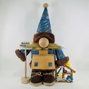 Puede incluir: Un gnomo azul y marrón que lleva un sombrero con estampado de herramientas y un delantal marrón. El gnomo sostiene un cartel que dice "Builders Gnome Biz" y tiene una pequeña caja de herramientas de madera con herramientas al lado.