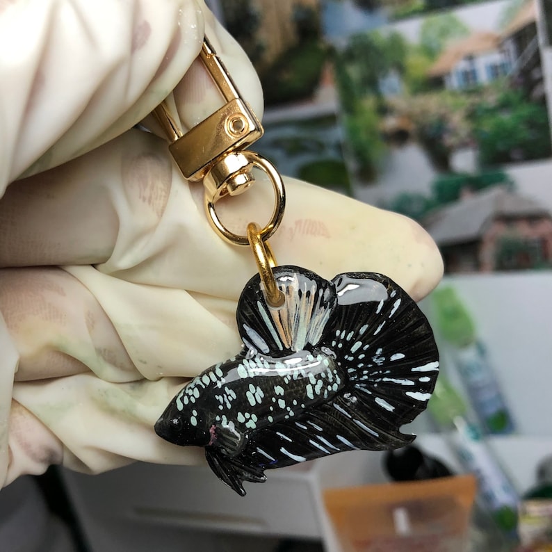 May include: A black and white betta fish keychain with a gold chain and clasp. The fish is detailed with white spots and fins.