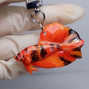 May include: Orange and black betta fish keychain made of polymer clay. The fish has a long, flowing tail and is detailed with black stripes and white spots.