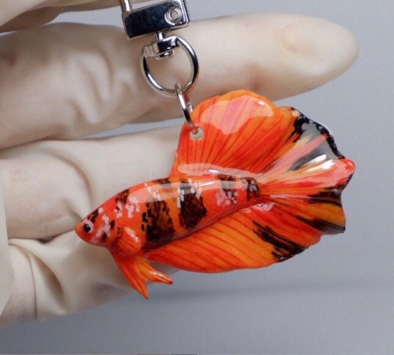 May include: Orange and black betta fish keychain made of polymer clay. The fish has a long, flowing tail and is detailed with black stripes and white spots.