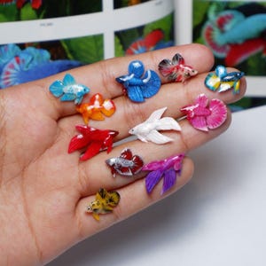 Peut inclure: Une collection de figurines miniatures de poissons betta colorés. Les figurines sont de différentes couleurs, notamment bleu, rouge, orange, blanc et violet. Chaque poisson a des nageoires détaillées et un motif unique. Les figurines sont présentées sur une main.