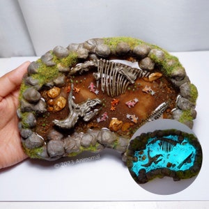 Op de afbeelding: Een miniatuur vijver van hars met een dinosaurus skelet, een triceratops schedel en kleine oranje vissen. De vijver is omgeven door rotsen en groen mos. De vijver gloeit blauw in het donker.