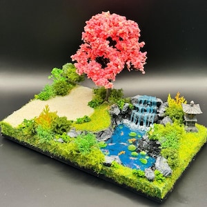 May include: A miniature landscape diorama featuring a vibrant pink cherry blossom tree, a small blue pond with orange koi fish and green lily pads, a cascading waterfall over grey rocks, a sandy path, green mossy terrain, and a stone pagoda.