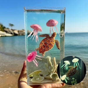 Puede incluir: Un bloque de resina transparente con una tortuga marina y medusas rosas suspendidas en un líquido azul, con coral y conchas en la base. El fondo muestra una escena de playa y océano.