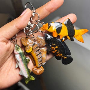 May include: A collection of colorful fish-shaped keychains. The keychains feature detailed designs of various fish species, including a rainbow trout, a goby, and others in shades of orange, black, yellow, and green. Each keychain is attached to a silver metal ring.