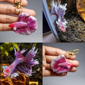 May include: A pink and white betta fish keychain with a gold chain. The fish is made of polymer clay and has a realistic appearance.