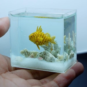 Op de afbeelding: Een miniatuur, helder acryl aquarium met een gedetailleerde gele vis met een textuurpatroon. De tank bevat koraal en een zandbodem, waardoor een kleine onderwaterwereld ontstaat. Het aquarium wordt in een hand gehouden.