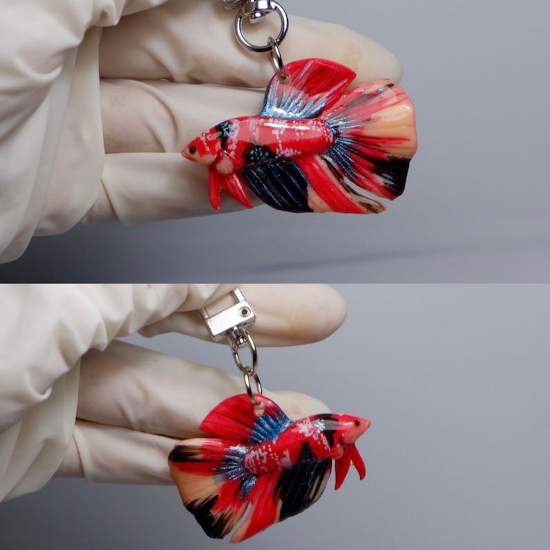 May include: A red, black, and white betta fish keychain. The fish is made of polymer clay and has a detailed, realistic design. The keychain is attached to a silver chain.