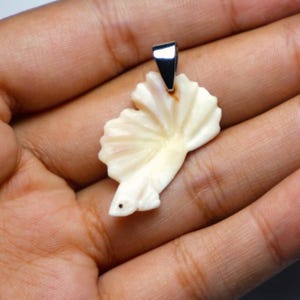 May include: A cream-coloured pendant in the shape of a fish with a fan-like tail. The pendant has a small, dark eye and a silver-coloured bail. The fish pendant is likely made of carved bone or a similar material.