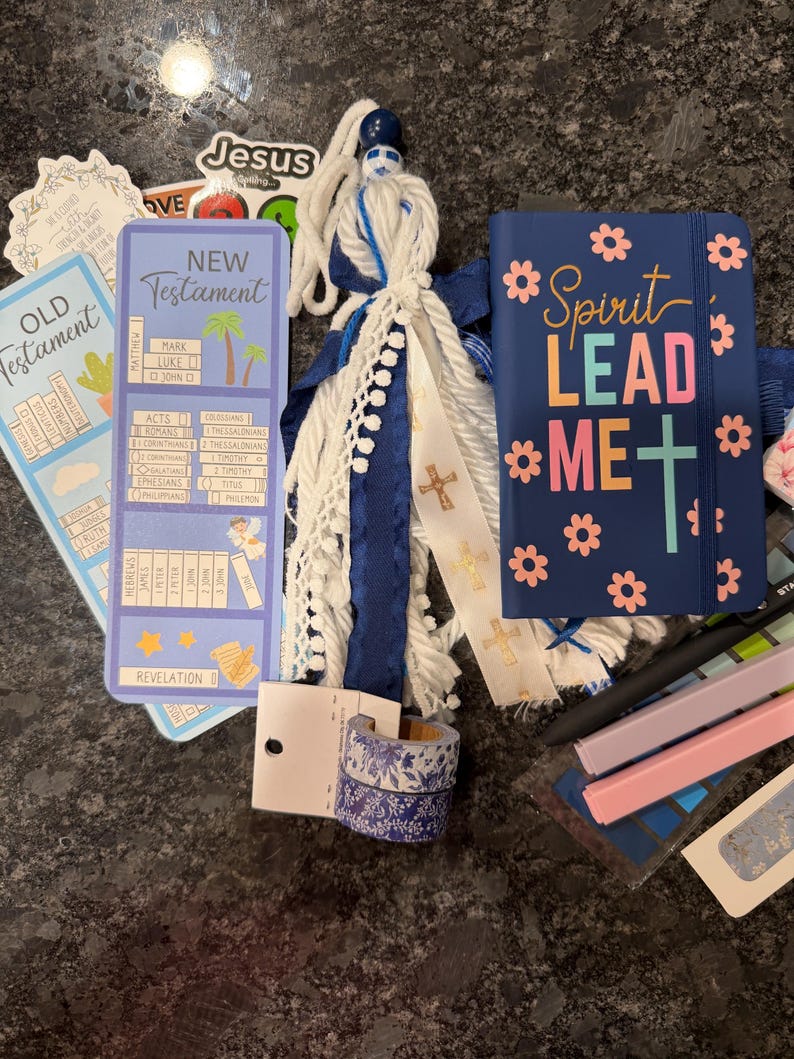 K&ouml;nnte beinhalten: Eine Sammlung von Gegenst&auml;nden, darunter ein blaues Notizbuch mit dem Text Spirit Lead Me, ein Lesezeichen f&uuml;r das Neue Testament, eine Quaste mit wei&szlig;en und blauen B&auml;ndern, zwei Rollen florales Washi-Tape und Aufkleber mit religi&ouml;sem Text.