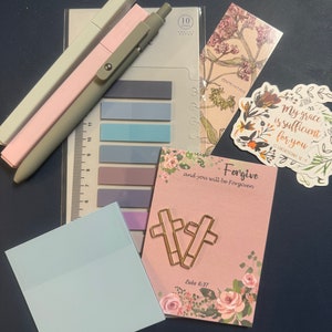 Puede incluir: Un bolígrafo rosa y gris, un juego de notas adhesivas de color pastel, una nota adhesiva azul, una tarjeta rosa con el texto "Forgive and you will be forgiven" y un clip de papel en forma de cruz dorada, un marcapáginas floral y una pegatina con el texto "My grace is sufficient for you 2 Corinthians 12:9"