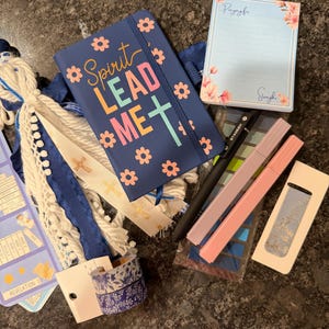 K&ouml;nnte beinhalten: Ein marineblaues Notizbuch mit dem Text Spirit Lead Me und Blumenmotiven, ein Notizblock mit Blumenrand, ein schwarzer Stift, zwei Pastellmarker, eine blau-wei&szlig;e Quaste, zwei Rollen gemustertes Washi-Tape und ein blaues Lesezeichen mit Goldtext.