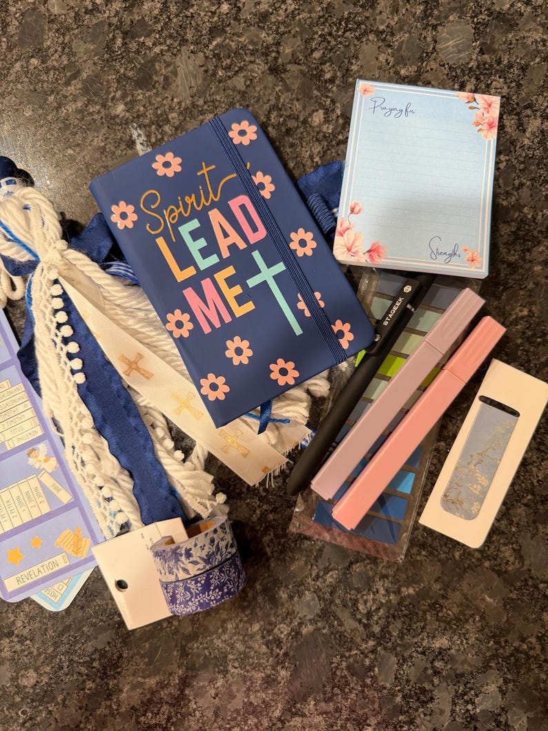 K&ouml;nnte beinhalten: Ein marineblaues Notizbuch mit dem Text Spirit Lead Me und Blumenmotiven, ein Notizblock mit Blumenrand, ein schwarzer Stift, zwei Pastellmarker, eine blau-wei&szlig;e Quaste, zwei Rollen gemustertes Washi-Tape und ein blaues Lesezeichen mit Goldtext.