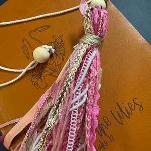 May include: A pink and white tassel with wooden beads and a white cord. The tassel is hanging from a brown leather journal with the words "The Lilies" and "Luke 12" engraved on the cover.