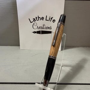 Pu&ograve; includere: Una penna artigianale con un fusto in legno venato e dettagli neri, esposta su un supporto trasparente. La penna presenta elementi argentati e una punta nera. Lo sfondo include una scheda bianca con il testo "Lathe Life Creations".