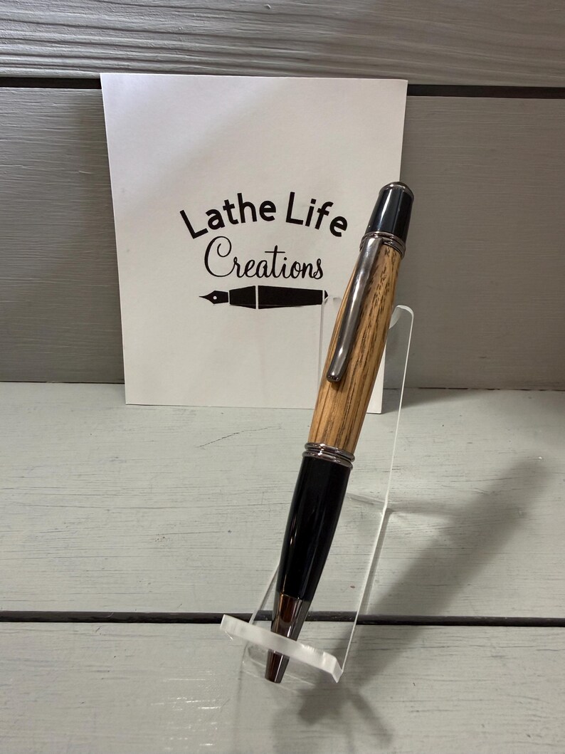 Pu&ograve; includere: Una penna artigianale con un fusto in legno venato e dettagli neri, esposta su un supporto trasparente. La penna presenta elementi argentati e una punta nera. Lo sfondo include una scheda bianca con il testo "Lathe Life Creations".