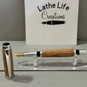 May include: A handcrafted fountain pen with a wooden barrel and silver accents. The pen features a gold nib and is displayed on a clear acrylic stand. The pen's cap is detached and placed next to the pen. The background includes a sign that reads "Lathe Life Creations."