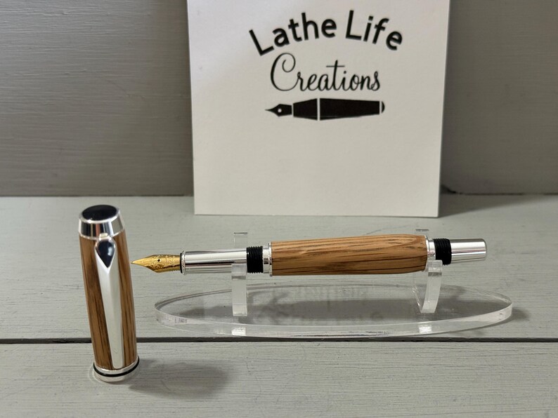 May include: A handcrafted fountain pen with a wooden barrel and silver accents. The pen features a gold nib and is displayed on a clear acrylic stand. The pen's cap is detached and placed next to the pen. The background includes a sign that reads "Lathe Life Creations."