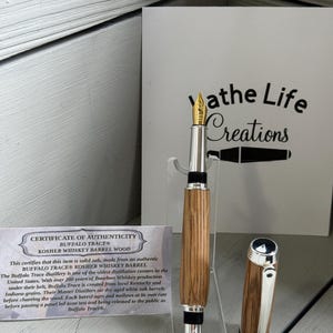 May include: A handcrafted fountain pen with a wooden barrel and silver accents. The pen features a gold nib and a matching cap. A certificate of authenticity is visible, along with the text "athe Life Creations".