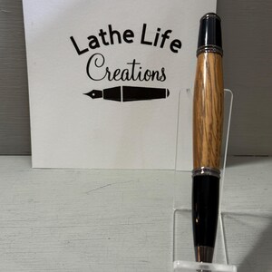 Pu&ograve; includere: Una penna in legno fatta a mano con punta e cappuccio neri, esposta su un supporto in acrilico trasparente. La penna presenta un motivo a venature del legno marrone chiaro. Una carta bianca dietro la penna recita "Lathe Life Creations".