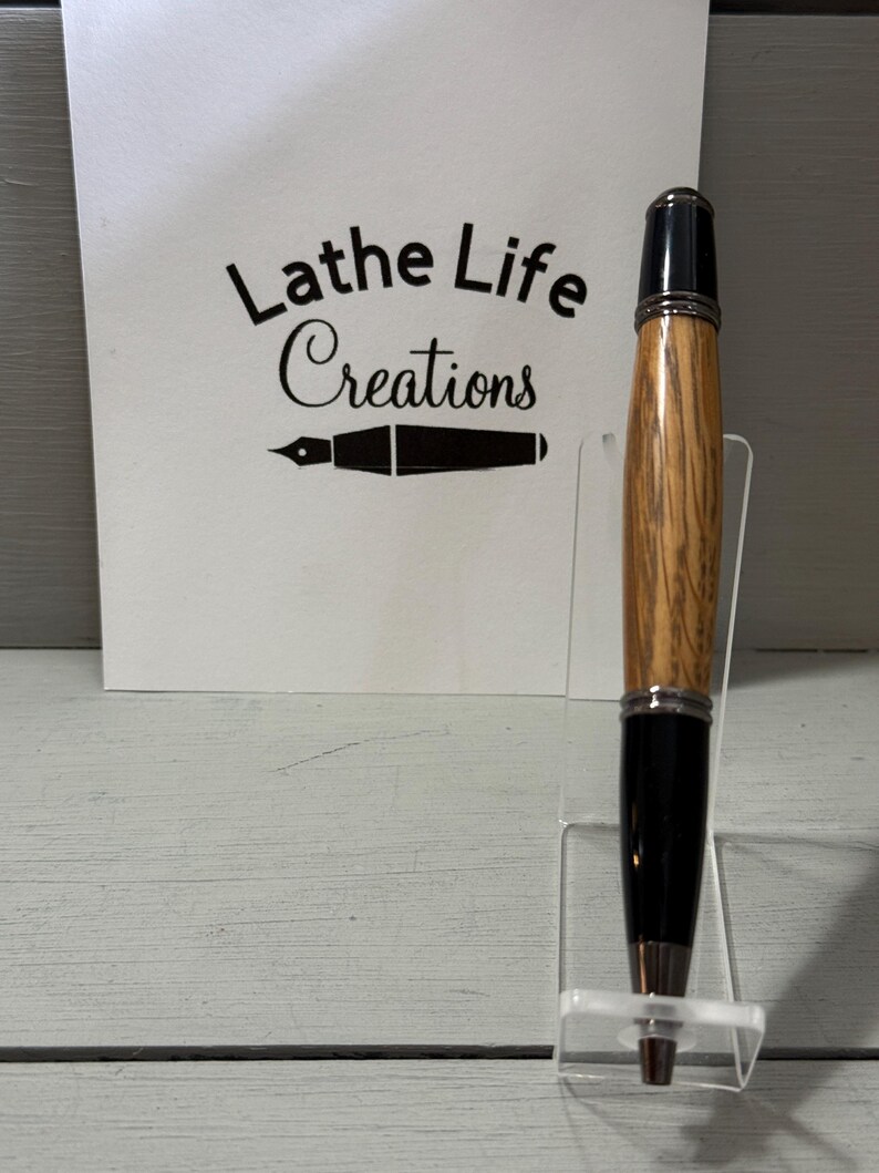Pu&ograve; includere: Una penna in legno fatta a mano con punta e cappuccio neri, esposta su un supporto in acrilico trasparente. La penna presenta un motivo a venature del legno marrone chiaro. Una carta bianca dietro la penna recita "Lathe Life Creations".