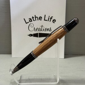Pu&ograve; includere: Una penna artigianale con fusto in legno e dettagli neri. La penna &egrave; esposta su un supporto in acrilico trasparente. Il testo "Lathe Life Creations" &egrave; visibile sullo sfondo.