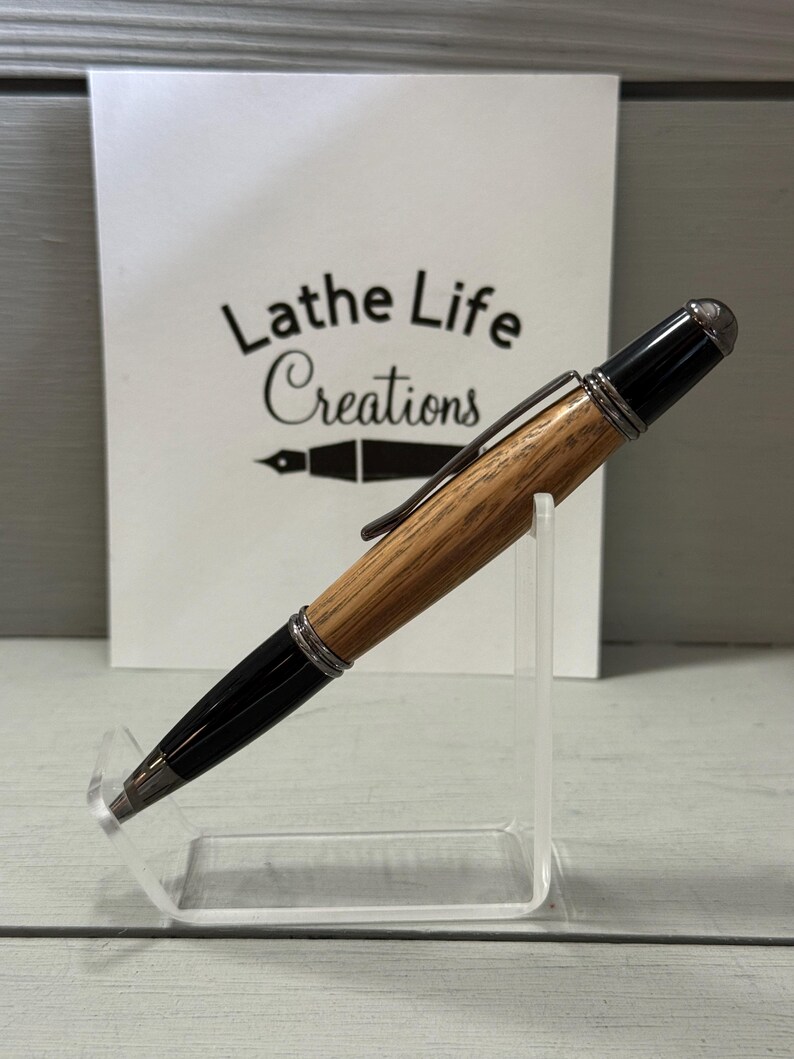 Pu&ograve; includere: Una penna artigianale con fusto in legno e dettagli neri. La penna &egrave; esposta su un supporto in acrilico trasparente. Il testo "Lathe Life Creations" &egrave; visibile sullo sfondo.