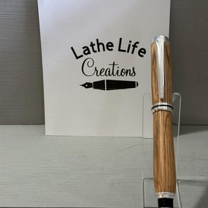 May include: A handcrafted wooden pen with silver accents, displayed on a clear acrylic stand. The pen features a natural wood grain pattern and a shiny metal tip. A white card behind the pen reads "Lathe Life Creations".
