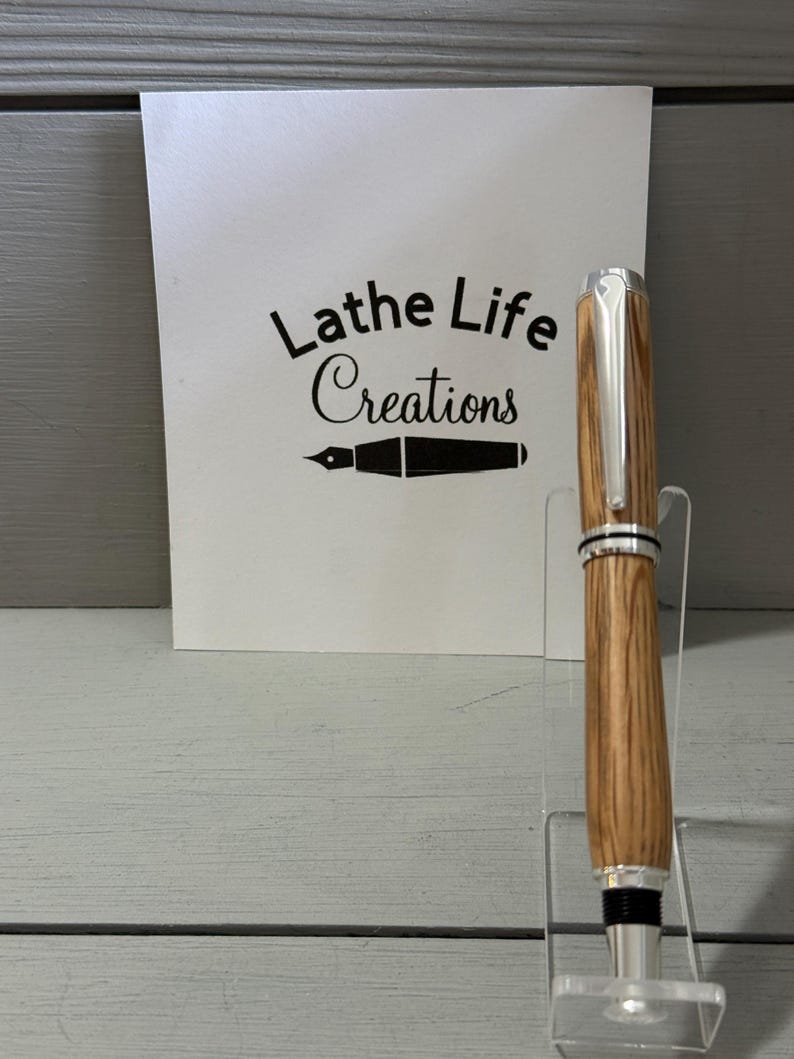 May include: A handcrafted wooden pen with silver accents, displayed on a clear acrylic stand. The pen features a natural wood grain pattern and a shiny metal tip. A white card behind the pen reads "Lathe Life Creations".
