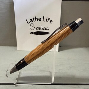 Puede incluir: Un portaminas de madera con acabado marr&oacute;n claro y detalles en negro. El l&aacute;piz tiene un clip plateado y una goma de borrar blanca. El fondo incluye una tarjeta blanca con el texto "Lathe Life Creations".