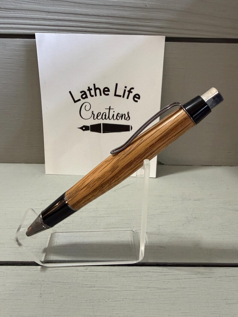 Puede incluir: Un portaminas de madera con acabado marr&oacute;n claro y detalles en negro. El l&aacute;piz tiene un clip plateado y una goma de borrar blanca. El fondo incluye una tarjeta blanca con el texto "Lathe Life Creations".