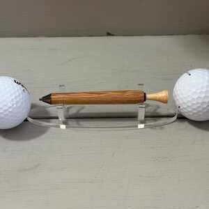 Puede incluir: Un bol&iacute;grafo de madera con un cuerpo marr&oacute;n claro y una punta de color claro, exhibido en un soporte acr&iacute;lico transparente. Dos pelotas de golf blancas est&aacute;n colocadas a cada lado del bol&iacute;grafo. Un regalo &uacute;nico.