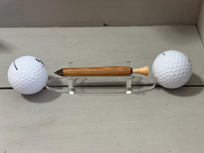 Puede incluir: Un bol&iacute;grafo de madera con un cuerpo marr&oacute;n claro y una punta de color claro, exhibido en un soporte acr&iacute;lico transparente. Dos pelotas de golf blancas est&aacute;n colocadas a cada lado del bol&iacute;grafo. Un regalo &uacute;nico.