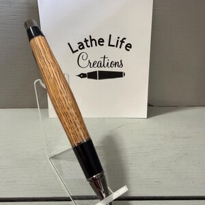 Puede incluir: Un bol&iacute;grafo artesanal con un cuerpo de madera marr&oacute;n claro y detalles en negro. El bol&iacute;grafo se exhibe en un soporte de acr&iacute;lico transparente. Una tarjeta blanca detr&aacute;s del bol&iacute;grafo dice "Lathe Life Creations".
