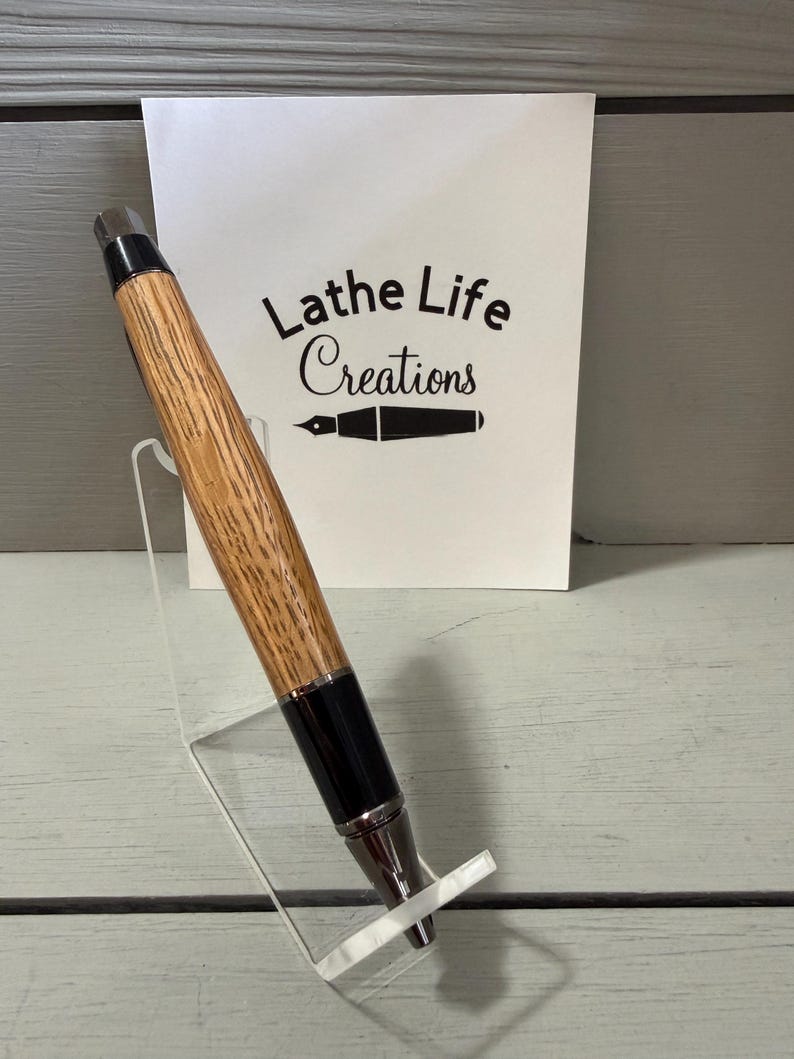Puede incluir: Un bol&iacute;grafo artesanal con un cuerpo de madera marr&oacute;n claro y detalles en negro. El bol&iacute;grafo se exhibe en un soporte de acr&iacute;lico transparente. Una tarjeta blanca detr&aacute;s del bol&iacute;grafo dice "Lathe Life Creations".