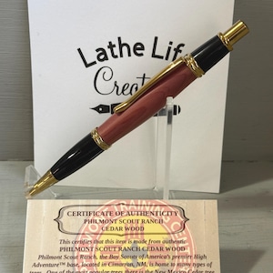 May include: A handcrafted pen made from cedar wood, featuring a gold-colored clip and accents. The pen has a black tip and end, with a reddish-brown wooden barrel. The pen is displayed with a certificate of authenticity from Philmont Scout Ranch.
