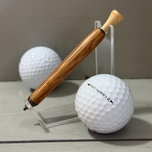Puede incluir: Un bol&iacute;grafo de golf de madera con una parte superior de tee de golf descansa sobre un soporte acr&iacute;lico transparente junto a una pelota de golf blanca. El bol&iacute;grafo tiene un acabado marr&oacute;n claro y un acento negro. Una segunda pelota de golf est&aacute; en el fondo.