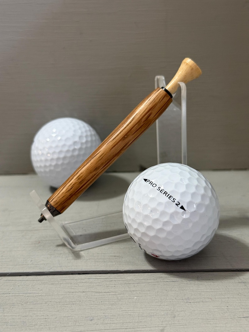 Puede incluir: Un bol&iacute;grafo de golf de madera con una parte superior de tee de golf descansa sobre un soporte acr&iacute;lico transparente junto a una pelota de golf blanca. El bol&iacute;grafo tiene un acabado marr&oacute;n claro y un acento negro. Una segunda pelota de golf est&aacute; en el fondo.