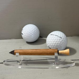 Puede incluir: Un bol&iacute;grafo de madera con acabado marr&oacute;n claro, exhibido en un soporte de acr&iacute;lico transparente. Dos pelotas de golf blancas est&aacute;n en el fondo. El bol&iacute;grafo tiene una punta oscura y un extremo claro.