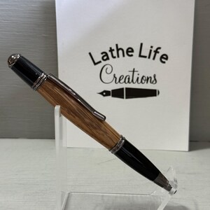 Pu&ograve; includere: Una penna artigianale con fusto in legno e dettagli neri, esposta su un supporto trasparente. La penna presenta finiture metalliche color argento e una punta nera. Lo sfondo include una scheda bianca con il testo "Lathe Life Creations".