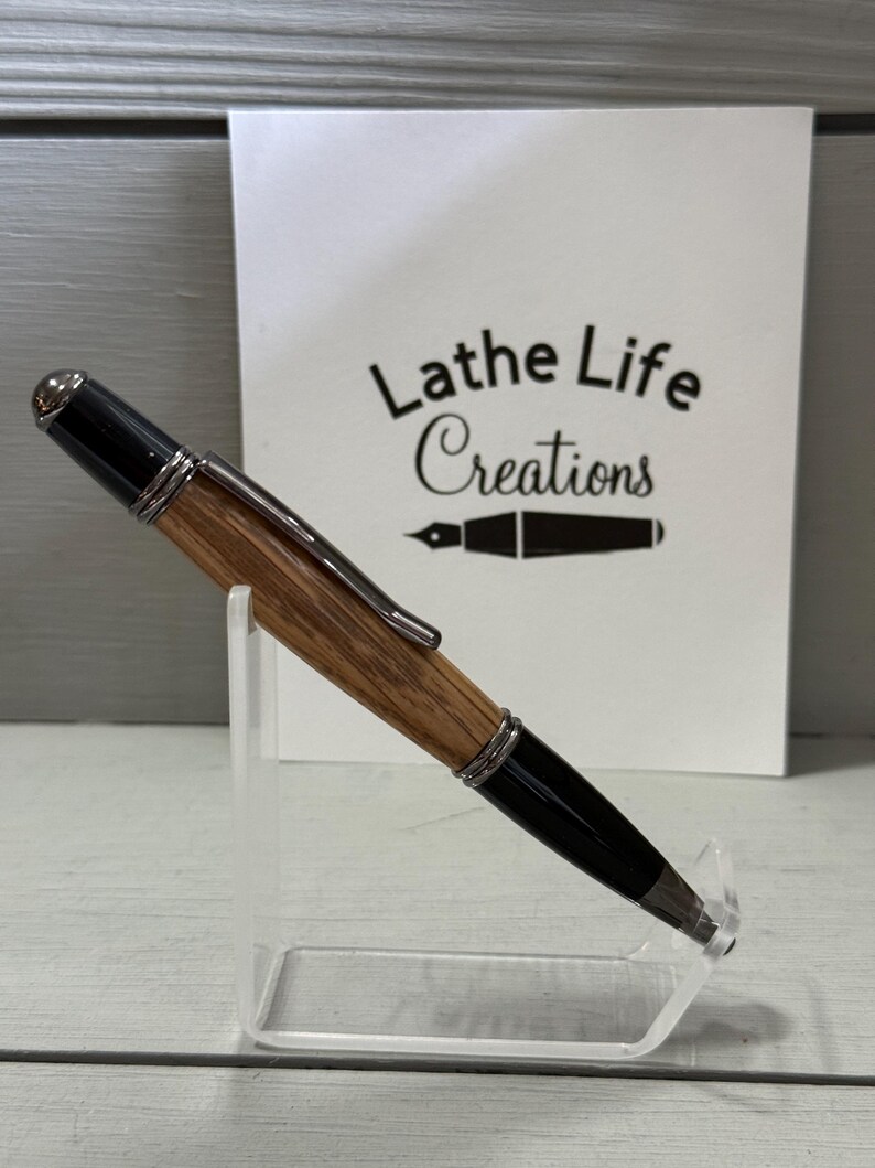 Pu&ograve; includere: Una penna artigianale con fusto in legno e dettagli neri, esposta su un supporto trasparente. La penna presenta finiture metalliche color argento e una punta nera. Lo sfondo include una scheda bianca con il testo "Lathe Life Creations".