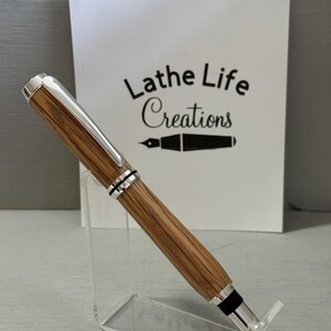 May include: A handcrafted pen with a wooden barrel and silver-colored accents. The pen is displayed on a clear acrylic stand. The text "Lathe Life Creations" is visible in the background.