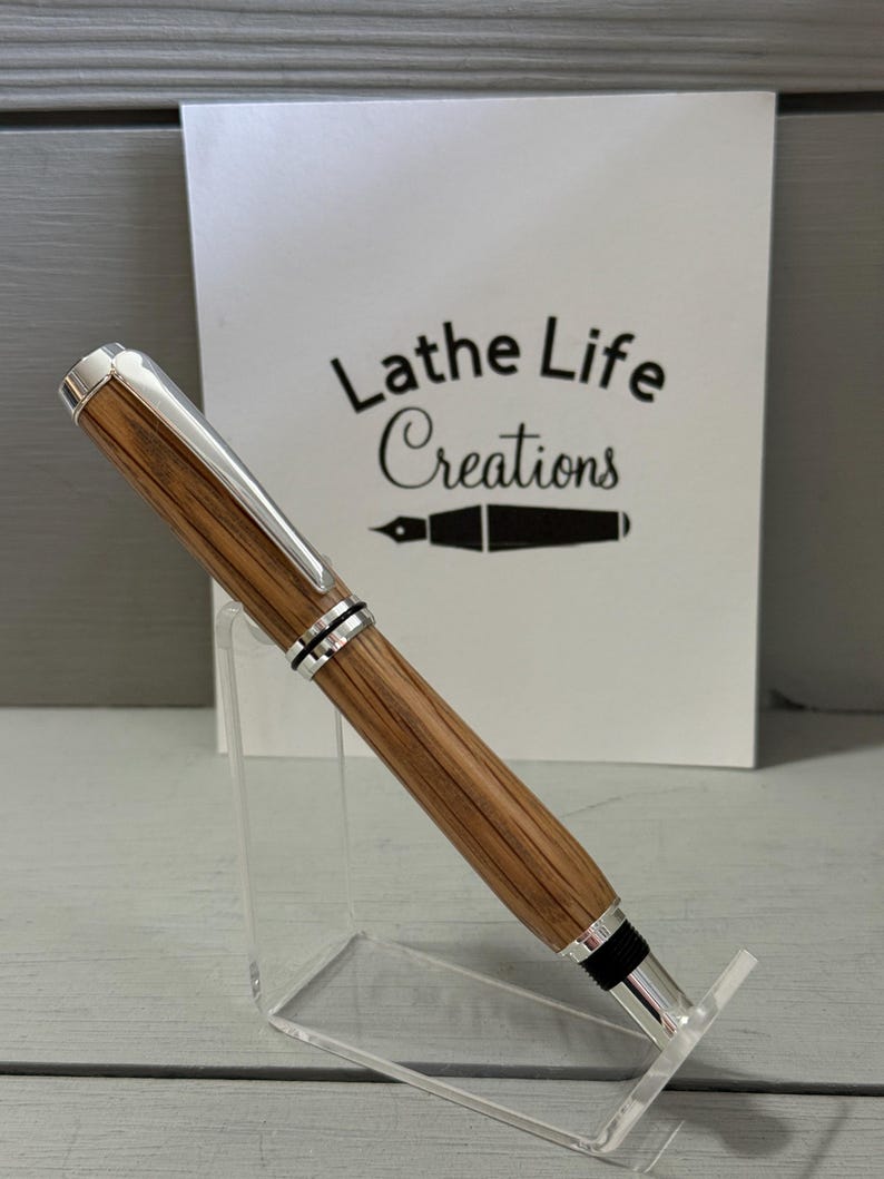 May include: A handcrafted pen with a wooden barrel and silver-colored accents. The pen is displayed on a clear acrylic stand. The text "Lathe Life Creations" is visible in the background.